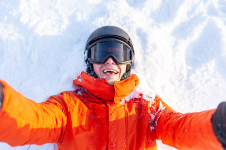 young cheerful guy in ski clothes lies on the snow in winter and takes selfie, man in orange jacket, helmet and glasses at the resort rejoices and takes photographsの写真素材