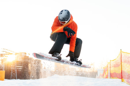 young cheerful guy in orange ski clothes jumps on snowboard in the mountains at resort, man in jacket, helmet and glasses does trick in the air and flies with snowboardの写真素材