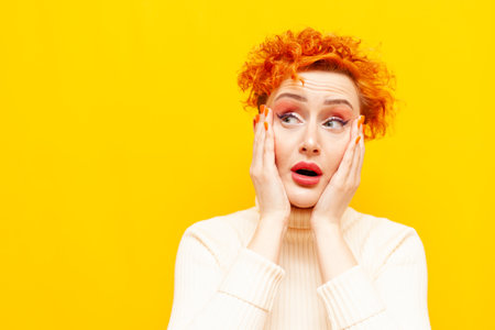 young red-haired excited woman with curly hair is surprised and looks away on yellow isolated background, shocked girl with beautiful make-up looks in amazement at the copy spaceの写真素材