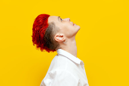 young short-haired red-haired girl looks up and thinks on a yellow isolated background, serious hipster woman with piercing raises her head and dreamsの写真素材