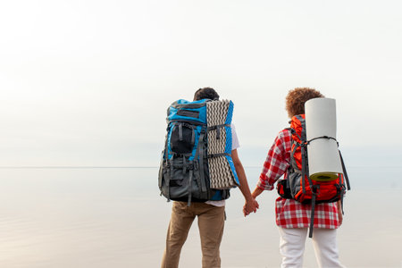young african american couple tourists with hiking equipment and backpacks standing on top of mountain and holding hands against sea and sky background, man and woman hiking with tents against lake and horizonの写真素材