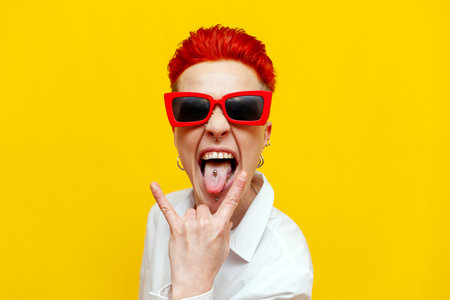 young short-haired red-haired punk girl shows rock gesture and screams on yellow isolated...