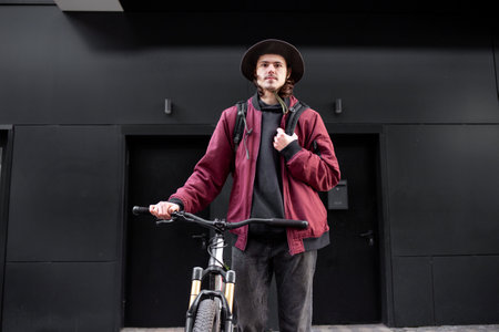 young student guy with backpack and bicycle stands against black wall and smiles, hipster man works...