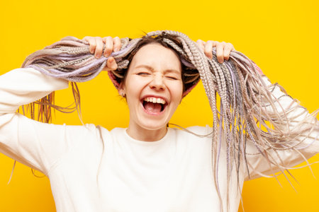 young girl with colored dreadlocks laughing and holding her hair over yellow isolated background,...