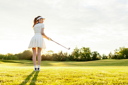 young attractive girl in sports uniform plays golf on field with lawn at sunset, woman with golf club plays sports outdoors in natureの写真素材