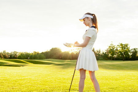 young girl in sports uniform plays golf on field with lawn at sunset, woman with golf club plays sports outdoors in natureの写真素材