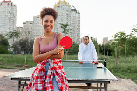 African American man and woman playing ping pong outdoors in the park, young woman standing near tennis table holding racket and looking at cameraの写真素材