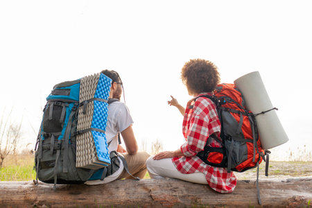 young couple with hiking equipment and backpacks sitting on tree in forest and showing route, man and woman resting with tents on camping site and pointing with handの写真素材