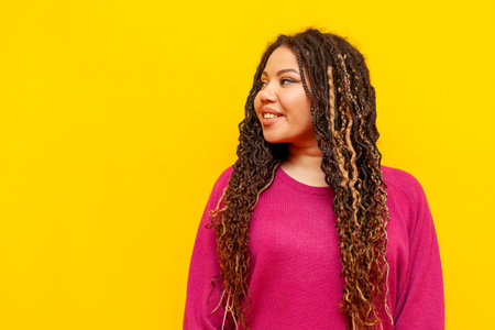 plump african american woman with dreadlocks smiles and looks away on a yellow isolated background, a big plus size girl advertises copy spaceの写真素材