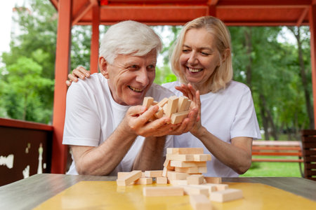 elderly couple of seniors in white t-shirts playing board game in park outdoors, old man and woman building a tower of wooden bricks and competing in game of balanceの写真素材