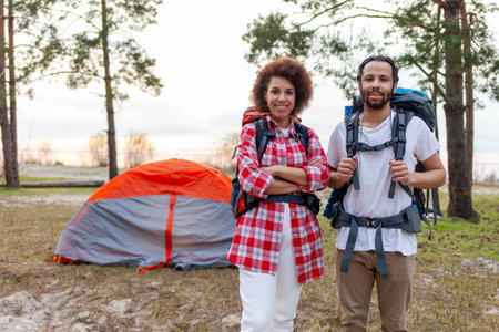 young african american couple tourists with hiking equipment and backpacks standing in forest against tent background, man and woman camping with tents and smilingの写真素材