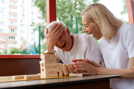 elderly senior couple playing board game and building a tower with wooden bricks outdoors in the park, old man shows facepalm and loses by making a mistakeの写真素材