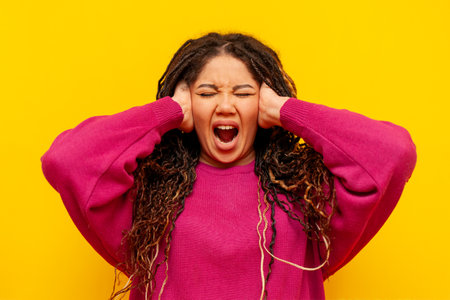 plump african american woman with pigtails screams and suffers from noise on yellow isolated background, plus size girl with dreadlocks covers her ears and ignoresの写真素材