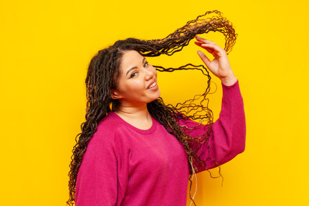 plump african american woman with dreadlocks smiling and waving her hair on yellow isolated background, big plus size girl dancingの写真素材