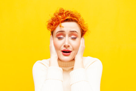 young red-haired woman with curly hair covers her ears on a yellow isolated background, a girl with a beautiful make-upの写真素材