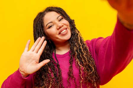 cheerful plump african american woman with pigtails takes selfie on smartphone and shows palm on yellow isolated background, big plus size girl with dreadlocks takes picture of herself and greetの写真素材