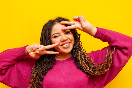 plump african american woman with dreadlocks smiling and showing peace gesture on yellow isolated background, plus size girl with pigtails showing two fingersの写真素材