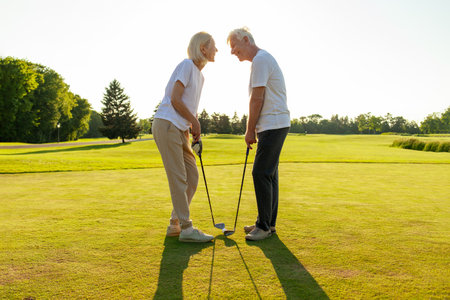 old senior couple in white clothes playing golf on golf course and chatting at sunset, elderly man and woman actively relaxing outdoors and laughing, pensioners on vacationの写真素材