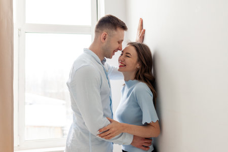 young happy couple in love stands near the window and laughs, the man hugs the woman and flirts at home and smilesの写真素材