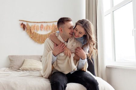 young happy couple in love hugging on the bed at home and smiling, the woman covers the man with a warm soft blanket and takes careの写真素材