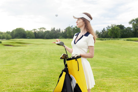 young attractive girl in sports uniform plays golf on field with lawn at sunset, woman with golf club plays sports outdoors in natureの写真素材
