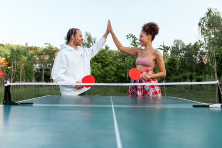 young african american couple playing table tennis outdoors in the park, man and woman team high fiving and celebrating victory and successの写真素材