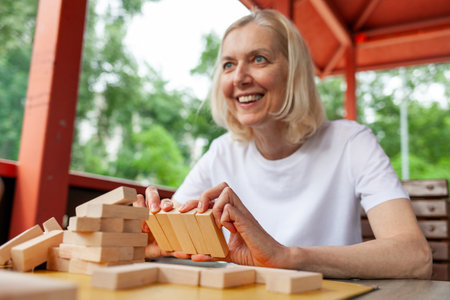 elderly couple of seniors in white t-shirts playing board game in park outdoors, old man and woman building a tower of wooden bricks and competing in a game of balanceの写真素材