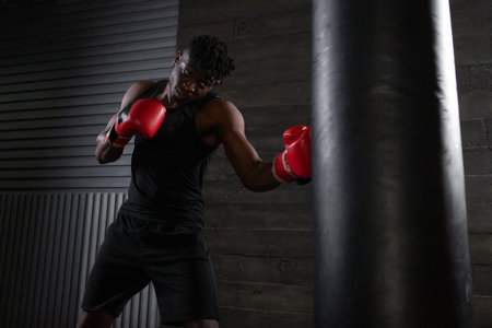 young strong african american boxer in red gloves hits punching bag, muscular athlete trains punch in dark gym and does sports on black backgroundの写真素材
