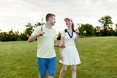 young sports couple playing golf together on field with lawn at sunset, woman practicing golf and talkingの写真素材
