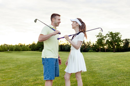 young couple playing golf together on field with lawn at sunset, woman practicing golf and talkingの写真素材