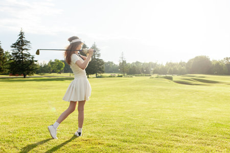 young attractive girl in sports uniform plays golf on field with lawn at sunset, woman with golf club plays sports outdoors in natureの写真素材