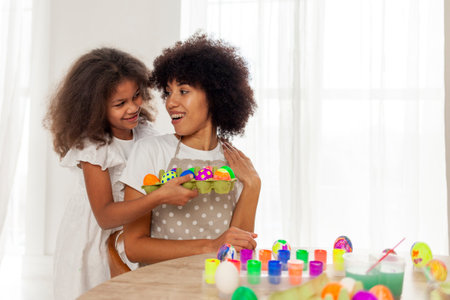 African American mother and daughter celebrate Easter and paint eggs with brush at home, woman with...