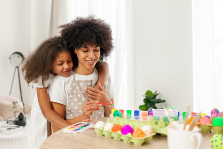 African American mother and daughter celebrate Easter and paint eggs with farbe and brush at home,...