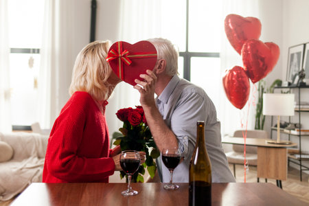 elderly senior couple celebrating valentines day at home and kissing, old man giving his wife gift, flowers and heart shaped balloons, retired people in love drinking wineの写真素材