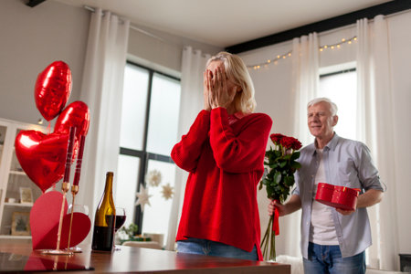 old man senior gives gift, flowers and heart-shaped balloons to his wife and congratulates on the holiday at home, elderly couple celebrates Valentines Day and makes romantic dateの写真素材