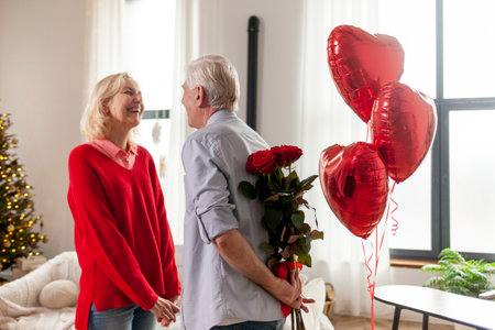 old man senior gives flowers and heart-shaped balloons to his wife and congratulates on the holiday at home, elderly couple celebrates Valentines Day and makes romantic dateの写真素材