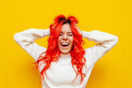 cheerful crazy red-haired woman screaming and holding her head with her hands on yellow isolated background, excited girl with colored curly hairstyle rejoicing and laughingの写真素材