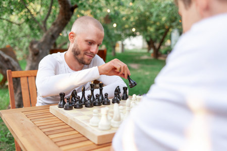 two young guys sitting at wooden table and playing chess outdoors in the park, male students competing in board game and planning strategyの写真素材