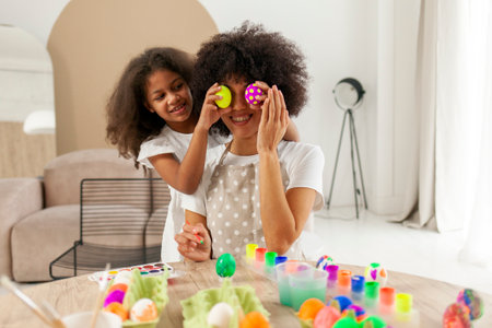 African American mother and daughter celebrate Easter and paint eggs with brush at home, woman with...