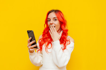shocked red-haired woman using smartphone and choosing online on yellow isolated background, excited...