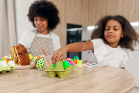 African American mother and daughter celebrate Easter and paint eggs with farbe and brush at home, woman with child decorate Easter eggsの写真素材
