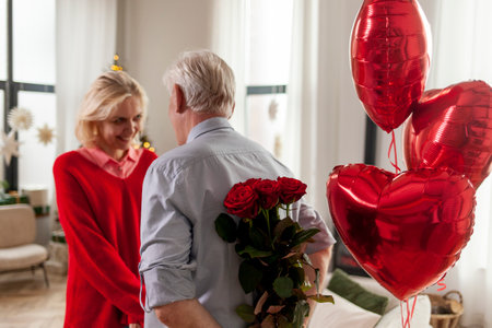 old man senior gives flowers and heart-shaped balloons to his wife and congratulates on the holiday at home, elderly couple celebrates Valentines Day and makes romantic dateの写真素材