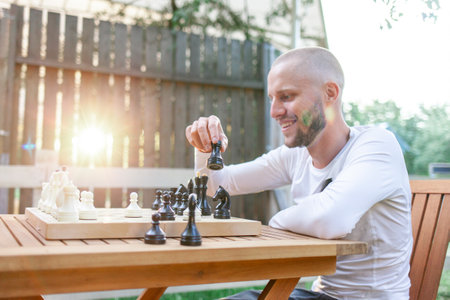 young cheerful guy plays chess outdoors in the park and smiles, the man makes move in the board gameの写真素材