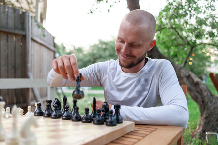 young cheerful guy plays chess outdoors in the park and smiles, the man makes move in the board gameの写真素材