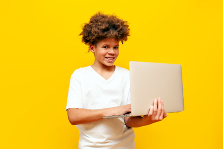 cheerful african american boy in white t-shirt using laptop on yellow isolated background and...