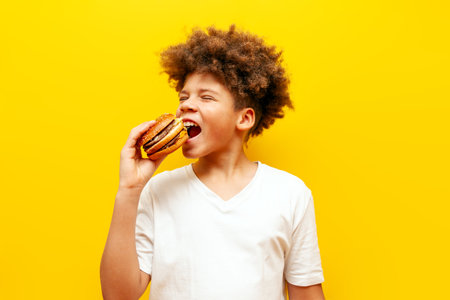 cheerful curly african american boy eating burger on yellow isolated background, hungry teenager holding cheeseburger and advertising fast foodの写真素材