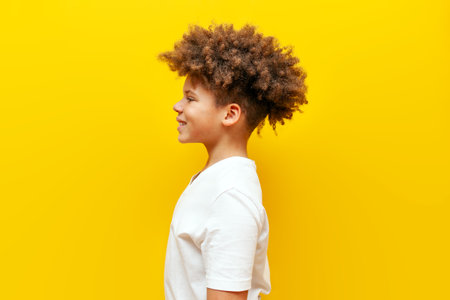 cheerful curly african american boy standing sideways and looking away on yellow isolated background, teenager in white t-shirt looking at copy space and smilingの写真素材