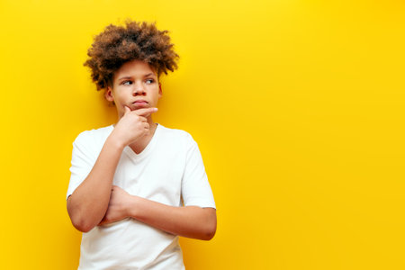 African American puzzled teenage boy in white t-shirt thinking and doubting on yellow isolated background, thoughtful insecure curly schoolboy kid planning and looking at copy spaceの写真素材