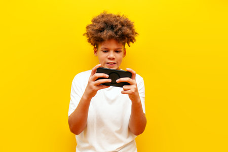 cheerful african american boy in white t-shirt playing video games on smartphone over yellow...