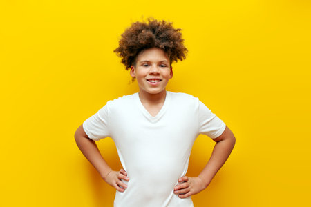 cheerful african american curly boy in white t-shirt standing on yellow isolated background, curly teenager smiling and looking at cameraの写真素材